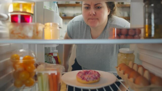 View from fridge of woman hesitating whether to eat donut or healthy food and choosing vegetables.