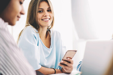 Portrait of young attractive blonde hipster girl enjoying spending free time with best friend talking during break in campus waiting for lesson beginning holding modern smartphone connected to wifi