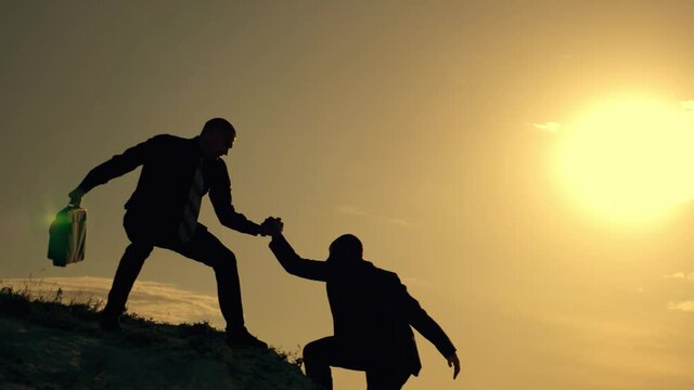 Businessmen climb to the top of the mountain, careers and one of them gives a helping hand. Silhouette of two men at sunset. Dressers help in crisis, overcoming obstacles. The concept of teamwork.