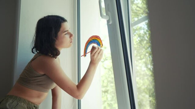 kid girl at home on the window draws a rainbow during coronavirus Covid-19 the quarantine period on self-isolation. Stay at home social media share. daughter lifestlye drawing rainbow child pandemic