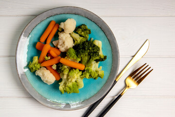 Steamed vegetables in a plate on a white wooden background, top view