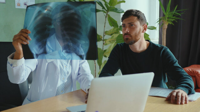 Beautiful black female doctor analyzing fluorography test of her patient reporting good results. Handsome bearded man is happy receiving positive news being very healthy.