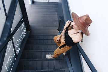 Obraz premium Fashionable brunette woman with long hair, wearing stylish large wicker hat, posing on the stairs. Young beautiful happy smiling fashionable traveller, tourist lady