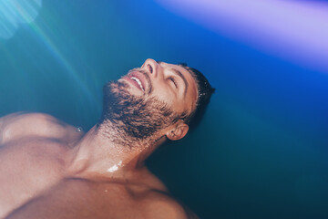 Handsome beard man floating in tank filled with dense salt water used in meditation, therapy, and alternative medicine.