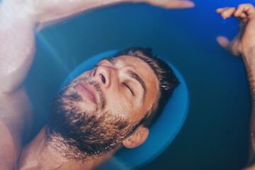 Handsome beard man floating in tank filled with dense salt water used in meditation, therapy, and alternative medicine.
