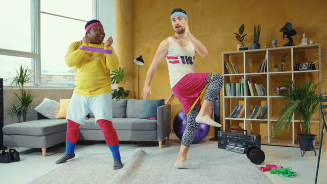 Multi-race friendly guys doing aerobics exercises at home together. Funny fake fitness jerks stretching wrong way with resistance bands having fun.