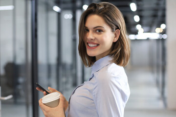 Businesswoman walking along the office corridor with paper cup and smartphone.