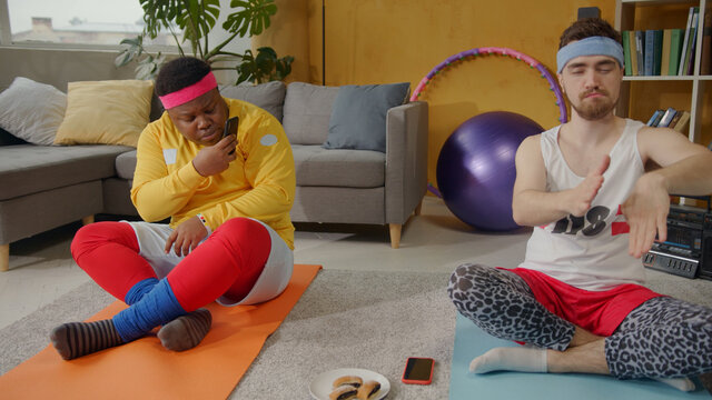 Comical scene. Black clumsy ridiculous guy eating cookies taking smartphone pictures of his concentrated funny friend meditating. Yoga at home. Sports parody.