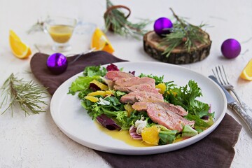 Warm salad with a mixture of salad leaves, baked duck and orange on a clay plate on a light concrete background in the New Year or Christmas style.