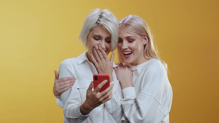 Two beautiful caucasian girlfriends LGBT couple family using mobile phone social media laughing having fun together at orange background. Copy space.