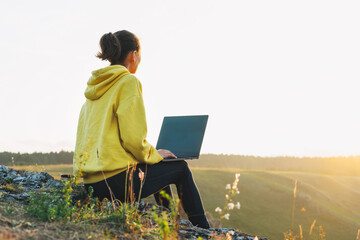 Young woman girl traveler freelancer in yellow hoodie with opened laptop looking at morning sunrise...