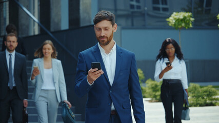 Attractive caucasian entrepreneur young businessman browsing in smartphone walking through crowd of busy corporate people outside business district. Nomophobia.