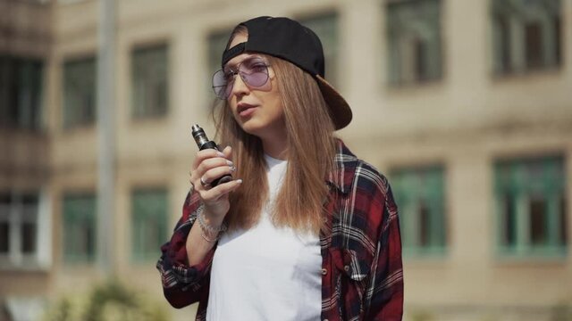 Pretty young hipster woman in black cap vape ecig. Girl vape popular ecig gadget, vaping device.Happy brunette vaper girl with e-cig. Portrait of smoker female model with electronic cigarette