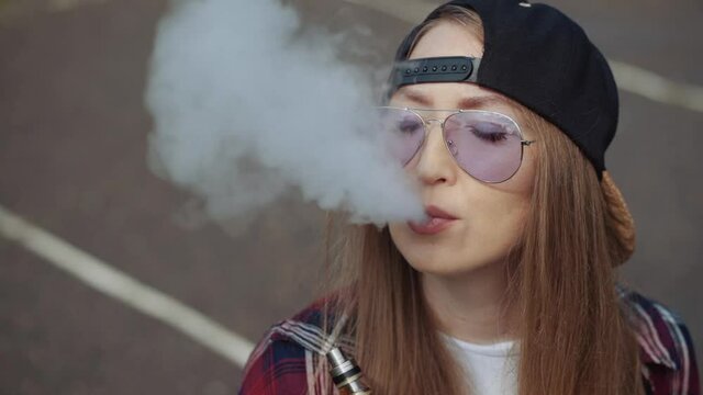 Close up pretty young hipster woman in black cap vape ecig. Girl vape popular ecig gadget, vaping device.Happy brunette vaper girl with e-cig. Portrait of smoker female model with electronic cigarette