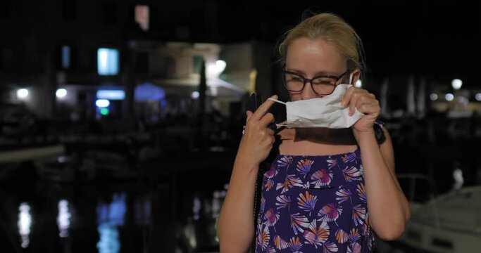 Evening shot of a young woman with glasses putting a protective mask on her face and taking a selfie with a cell phone. Summer evening on the island of Brač on the Croatian coast.
