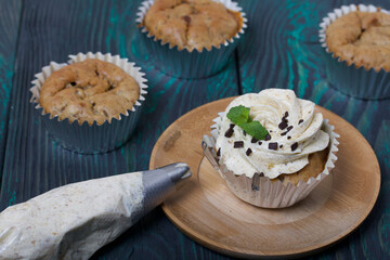 Banana cupcake. With butter cream and banana jam. Decorated with mint leaves and chocolate sprinkles. Next to them are cupcakes without cream and a pastry bag with cream.