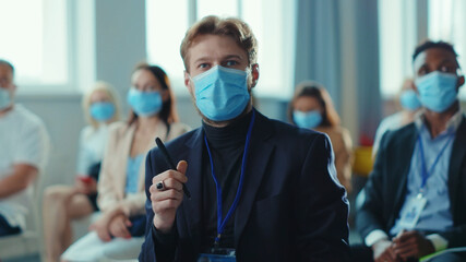 Young stylish start-up entrepreneur asking question on lecturer on modern conference meeting business seminar. Crowd of diverse people wearing masks. Coronavirus.