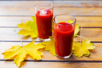 Tomato juice is poured into two glass glasses on a wooden table with yellow fallen maple leaves, in autumn. Selective focus.
