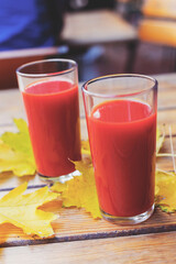 Tomato juice is poured into two glass glasses on a wooden table with yellow fallen maple leaves, in autumn. Selective focus. Tinted background