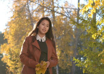 Portrait of a girl against the background of autumn trees. A girl in a leather jacket holds yellow autumn leaves.