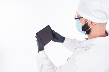 A doctor in a mask and rubber gloves. The doctor is holding an electronic tablet. Doctor in a white coat and cap on a light background.