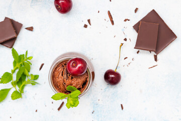 Homemade dark chocolate mousse garnish with sweet cherry and mint and pieces of chocolate on light blue background.