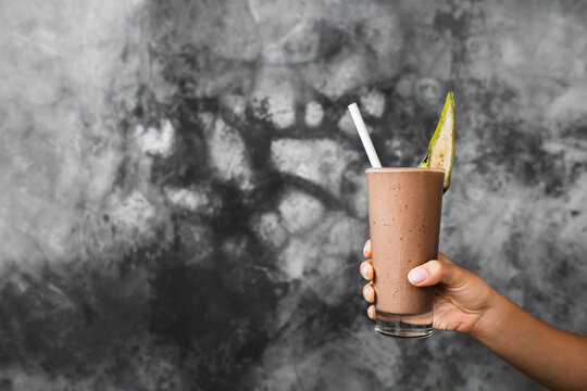 Glass Of Fresh Cold Chocolate Milk Shake In Woman Hands On Background Of Gray Concrete Cement Textured Wall. Cocoa Smoothie With Banana Piece As Decoration. Summer Concept.