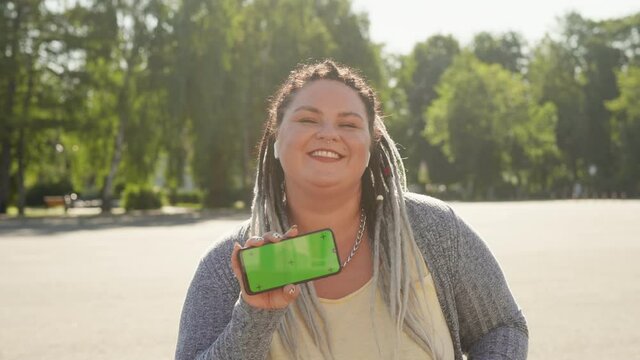 Beautiful stout girl with chromakey on her smartphone advertising mobile app. Commercial, copy space concept. Young pretty fat woman with dreads showing green screen phone to camera and dancing.