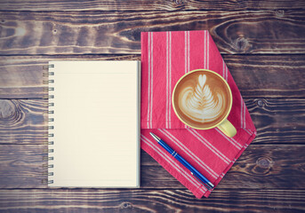 Top view, flat lay latte art coffee in a yellow cup with a notebook, a blue pen, and red fabric on the old wooden table background.