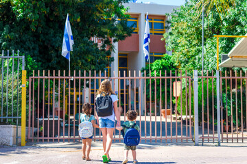 Happy diverse school children going to school outdoor at sunny day.Rear view of old sister and little girl and boy preschoolers with backpack go hand in hand to the kindergarten. Beginning of lessons.