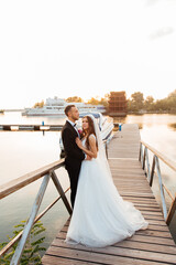Fototapeta na wymiar Beautiful luxurious couple, the groom in a black suit and the bride with a veil and a white wedding dress, holding hands, walk by the pool on the pier, next to the yacht and the ship