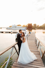 Beautiful luxurious couple, the groom in a black suit and the bride with a veil and a white wedding dress, holding hands, walk by the pool on the pier, next to the yacht and the ship