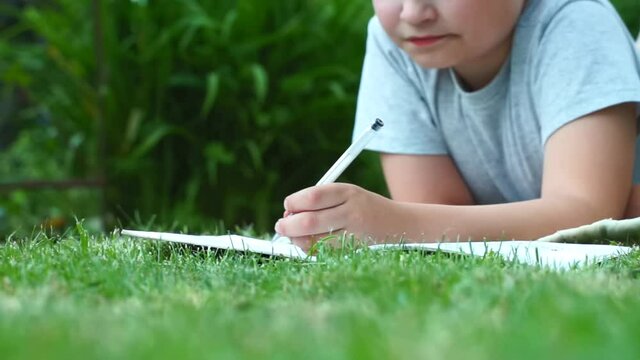 close up child hands writing thoughts and dreams in a diary book laying on grass at nature. dreamy child dreaming outdoors. Thoughtful schoolboy making notes in a notebook