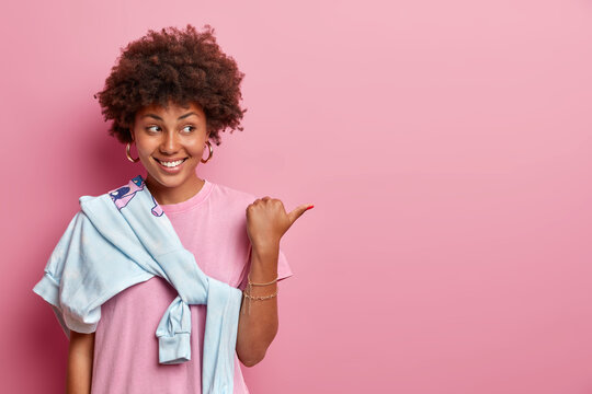 Pretty Girl With Afro Hair Helps To Pick Best Choice, Points Thumb Aside On Copy Space, Advertises Product, Smiles Happily, Wears Rosy T Shirt And Sweater Tied Over Shoulder. Your Promo Here