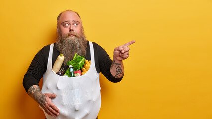Chubby bearded adult man eats unhealthy food, points finger aside on copy space, tells where he bought all these products, leads junk lifestyle, has obesity problem, isolated on yellow background