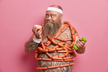 Never enough food. Fat greedy man cannot deny eating sweet appetizing doughnut, trains muscles with dumbbell, does sport exercises for loosing weight, many sausages around naked tattooed body