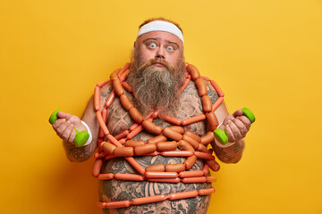 Problem of obesity. Shocked tired man with big tummy and thick beard, gets lots of energy from sausages, trains muscles, holds dumbbells, concerns about health, isolated on yellow background