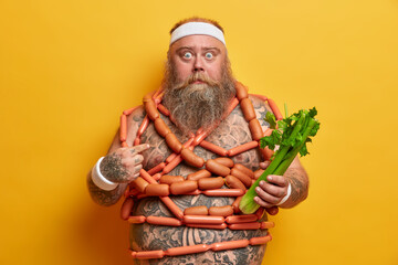 Shocked obese bearded man points at himself and looks with bugged eyes, holds celery and wrapped with sausages, chooses appetizing junk food over organic one, suffers from gluttony and overweight