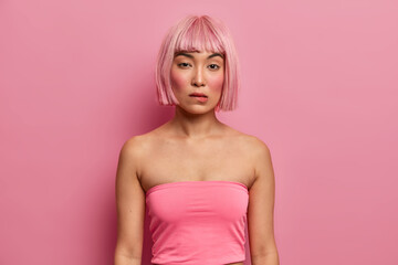 Serious adorable woman with eastern appearance, pink bob hairstyle, wears tank top, bites lips and looks directly at camera, thinks about good decision, has mysterious expression. Fashion girl