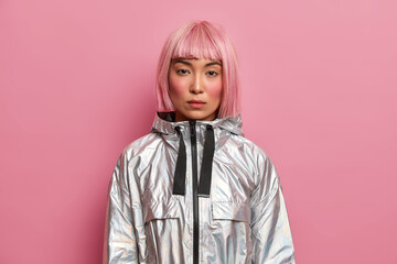 Portrait of serious woman with stylish pink hairstyle, perfect fresh clean skin, looks at camera with calm confident expression, dressed in silver jacket, poses over rosy background. Hair coloring
