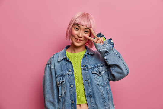 Photo of young alluring woman with trendy pink hairdo, shows victroy gesture over eye, wears stylish denim jacket, has fun, poses against rosy studio background, enjoys awesome adventure. Peace sign