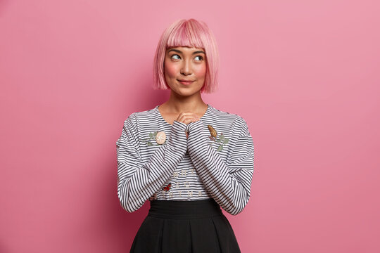 Thoughtful dreamy woman with pink hairstyle, keeps hands together, looks mysteriously, wears fashionable striped jumper and black skirt, pictures something beautiful in mind, has thought in bulb