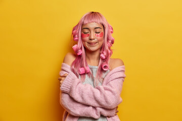 Dreamy pleasant looking woman with pink hair, hugs herself gently, enjoys softness of warm jumper, wears curlers, makes perfect hairstyle, wears patches under eyes for reducing wrinkles, stands indoor