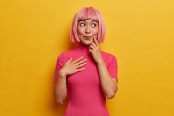 Photo of smiling happy Asian woman wears pink hair wig, imagines something pleasant, looks with dreamy peaceful expression, recalls good memories and makes wish, dressed in rosy casual clothes