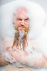 Close-up portrait of funny red-bearded bald man taking a bath with foam. Cheerful naughty guy at spa treatments. A parody of glamorous girls.