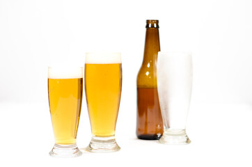 Two glasses full of beer with foam along with a brown glass bottle and another cold empty glass on a white background.