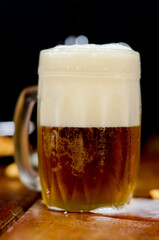 A mug of cold beer with foam in the foreground on a wooden table with an out of focus black background.