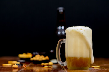 A cold beer mug with the foam falling down with beer bottles behind with some savory snacks on a wooden table with a black background.