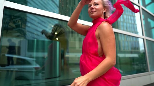 An woman with pink hair poses on the street during the day against the background of a city business center, her back leaning against the Windows. She is wearing a pink dress along with a scarf.