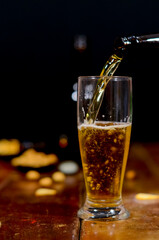 A beer bottle filling a glass in the foreground with a black bucket with beers inside, some salty snacks on a wooden table with a black background.